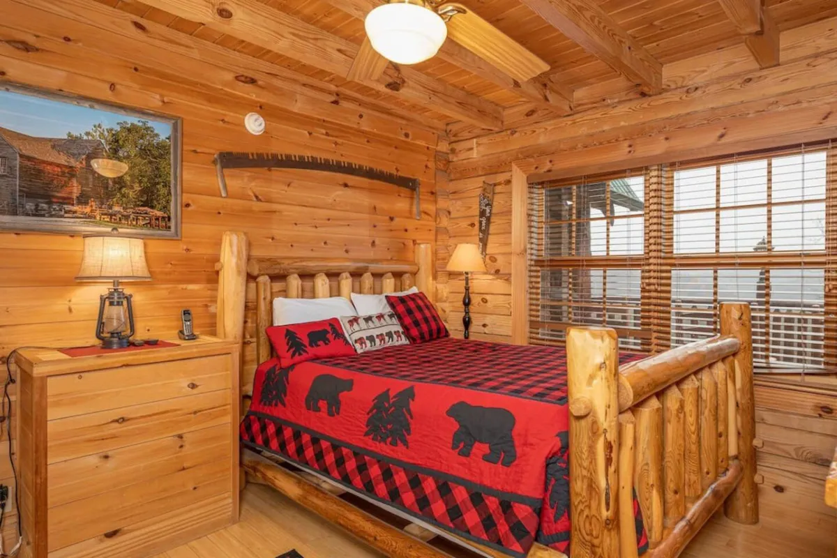 A warmly lit bedroom with a log bed covered in plaid bedding, complemented by wooden furniture, a framed artwork on the wall, and a table lamp on the nightstand. The window lets in soft natural light, enhancing the tranquil and cozy ambiance.