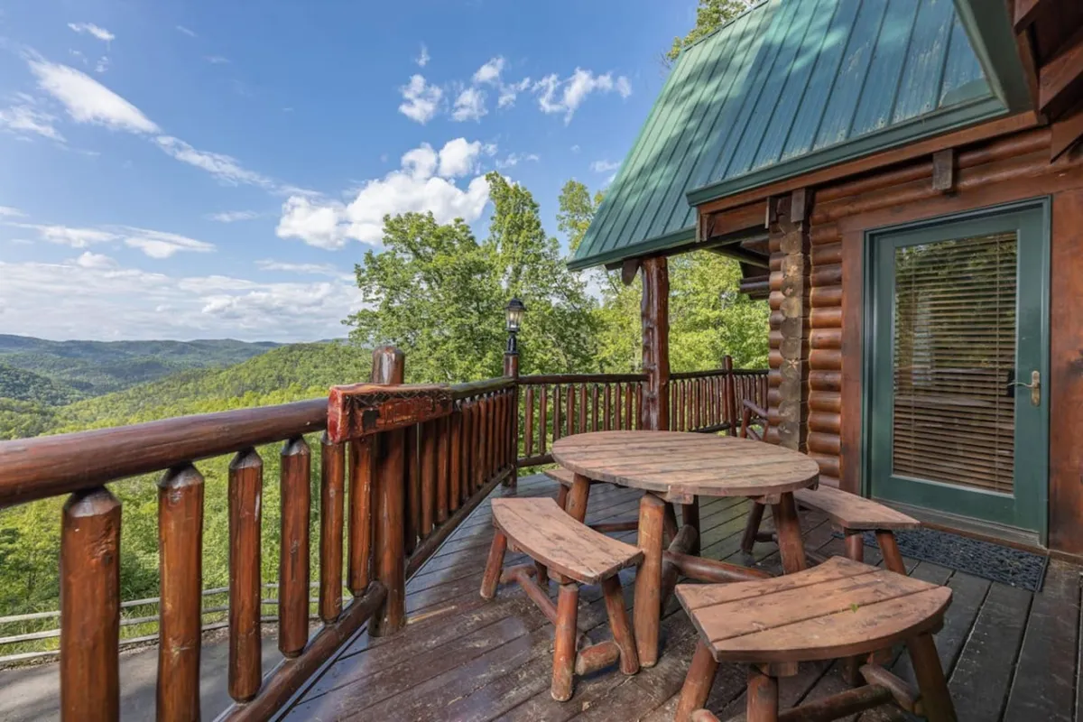 A scenic wooden deck furnished with a round table and surrounded by lush greenery, offering breathtaking views of the Smoky Mountains. The deck is ideal for rocking-chair relaxation, outdoor dining, or soaking in the landscape with friends and family.