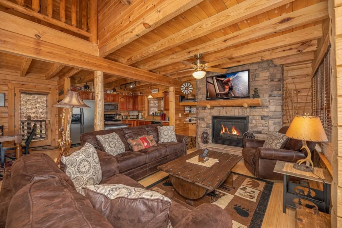 A warmly lit living room featuring plush leather sofas arranged around a stone fireplace, with exposed wooden beams overhead. The 75-inch TV is positioned above the mantel, and inviting décor creates a cozy, communal atmosphere ideal for group relaxation and entertainment.