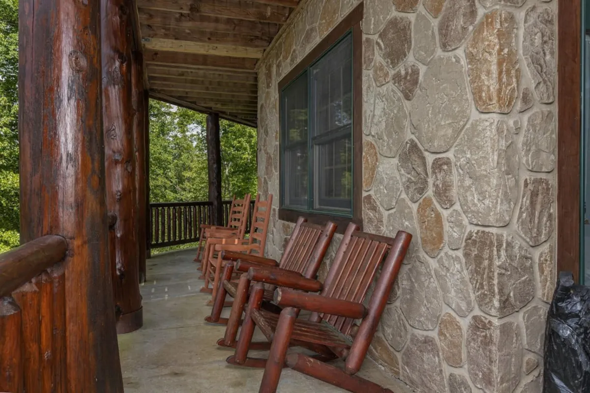A welcoming porch scene with rustic wooden rocking chairs, sturdy wooden pillars, a stone wall, and vibrant surrounding greenery. The setup suggests a space for relaxation, enjoying quiet moments, and soaking in the natural mountain atmosphere.