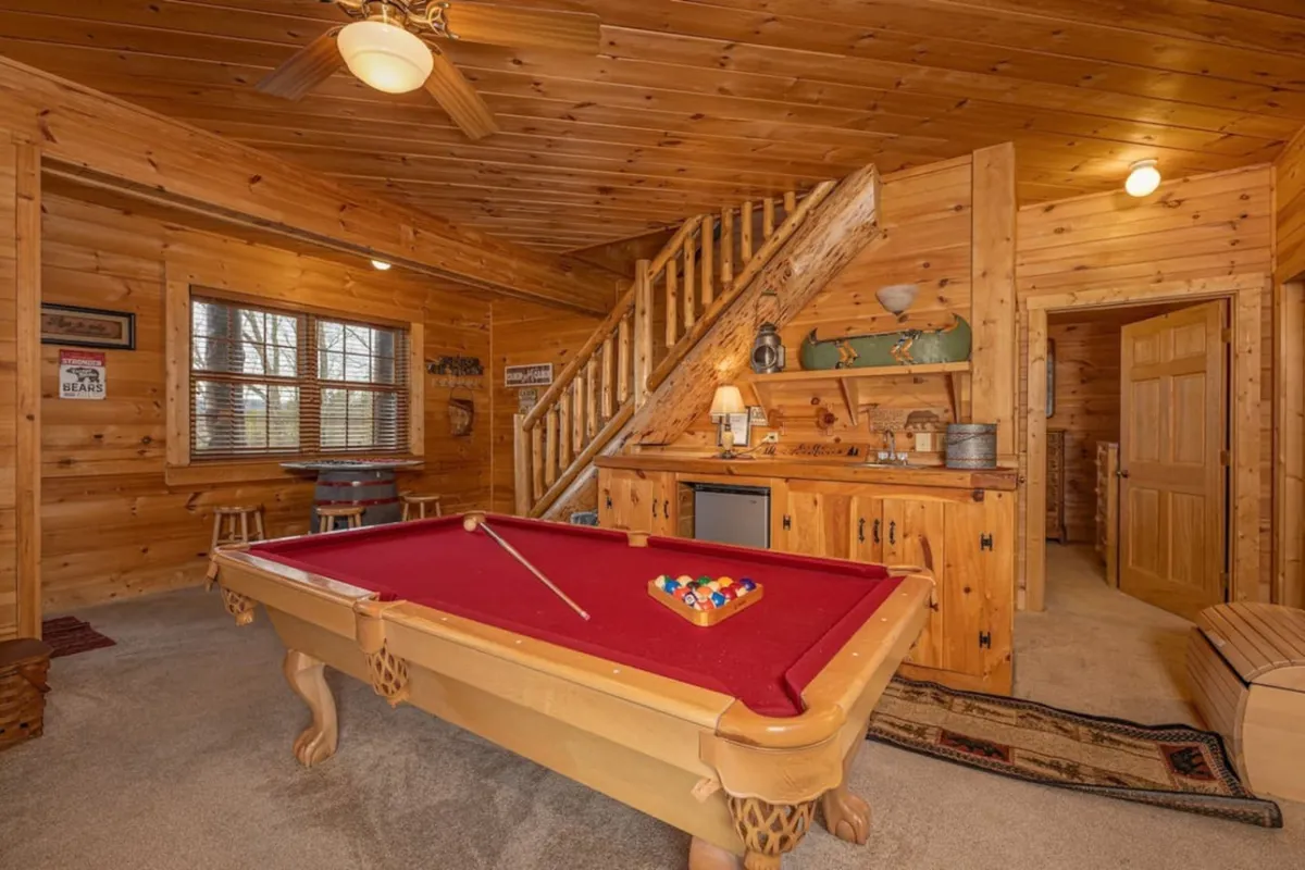 A spacious game room featuring a classic pool table prominently in the center, surrounded by rustic wooden walls and a staircase. A cozy bar area sits in the background, with inviting decor that sets the tone for lively gatherings and friendly competition.