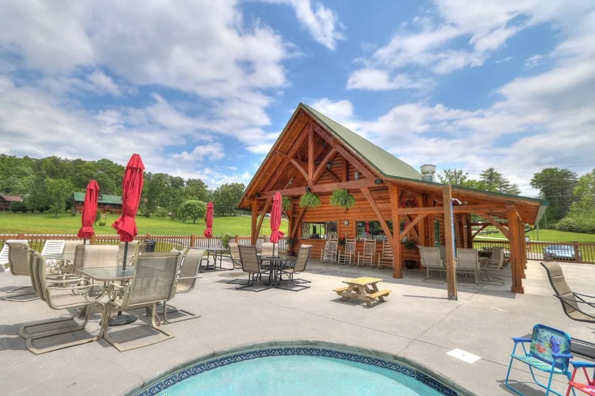 A vibrant pool area featuring a wooden pavilion, red umbrellas, and a lush green lawn. The scene conveys a cheerful, social atmosphere ideal for family gatherings, sunbathing, and summer barbecues.