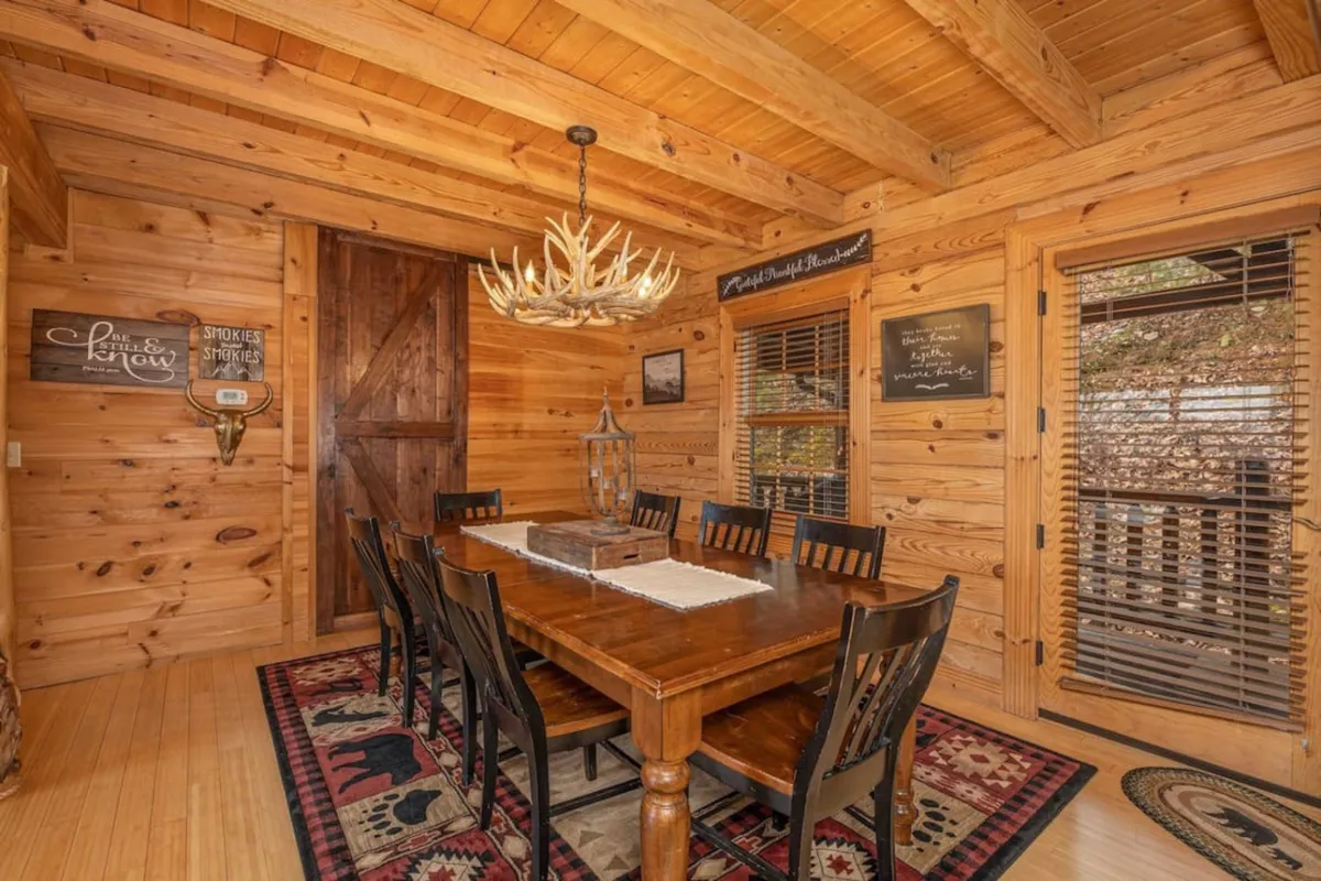 A large wooden dining table set beneath a rustic antler chandelier, surrounded by warm wooden walls and decorative touches. The expansive table allows for group meals, celebrations, and board games, embodying the essence of shared dining and gathering.