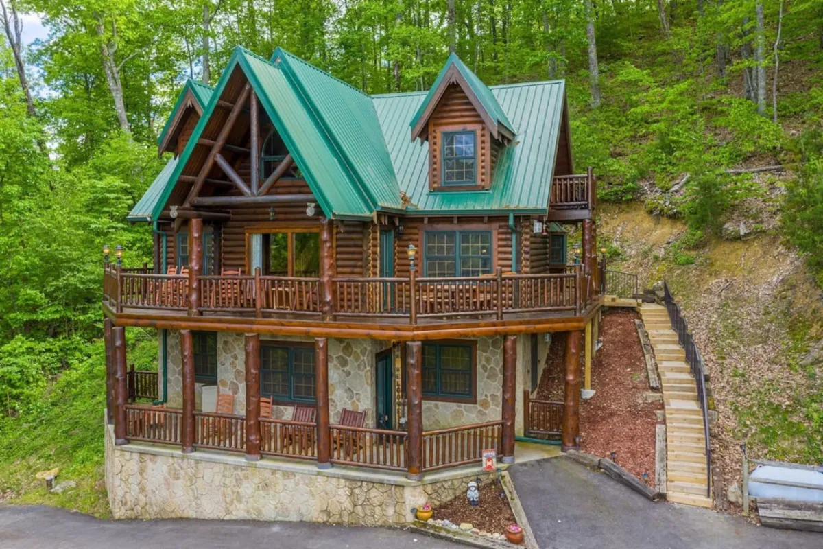 A welcoming log cabin with a green metal roof, set amidst lush greenery. The cabin features a stone facade and balconies, with a spacious porch perfect for outdoor gatherings and relaxation. The driveway and surrounding landscape set the scene for family arrivals and adventures.