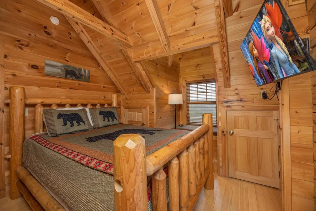 A bedroom with a log bed adorned in bedding with bear motifs, a flat-screen TV nearby, and wood-paneled walls. The space feels inviting and designed for relaxation, ideal for couples or families.
