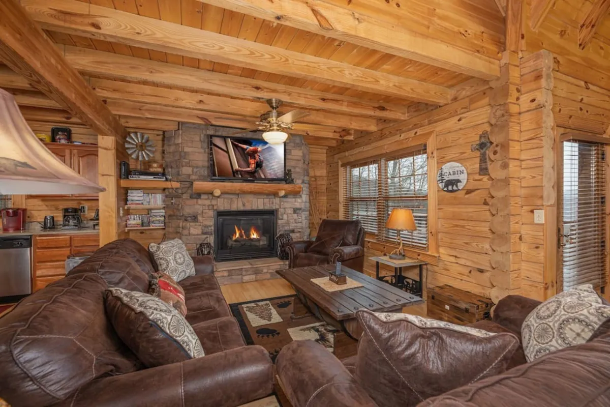 A cozy cabin living room with a glowing fireplace, large comfortable sofas, a coffee table, and a television. The windows let in natural light and showcase panoramic views, while the ambiance is warm and inviting—ideal for fireside chats, movie nights, or relaxing after outdoor adventures.