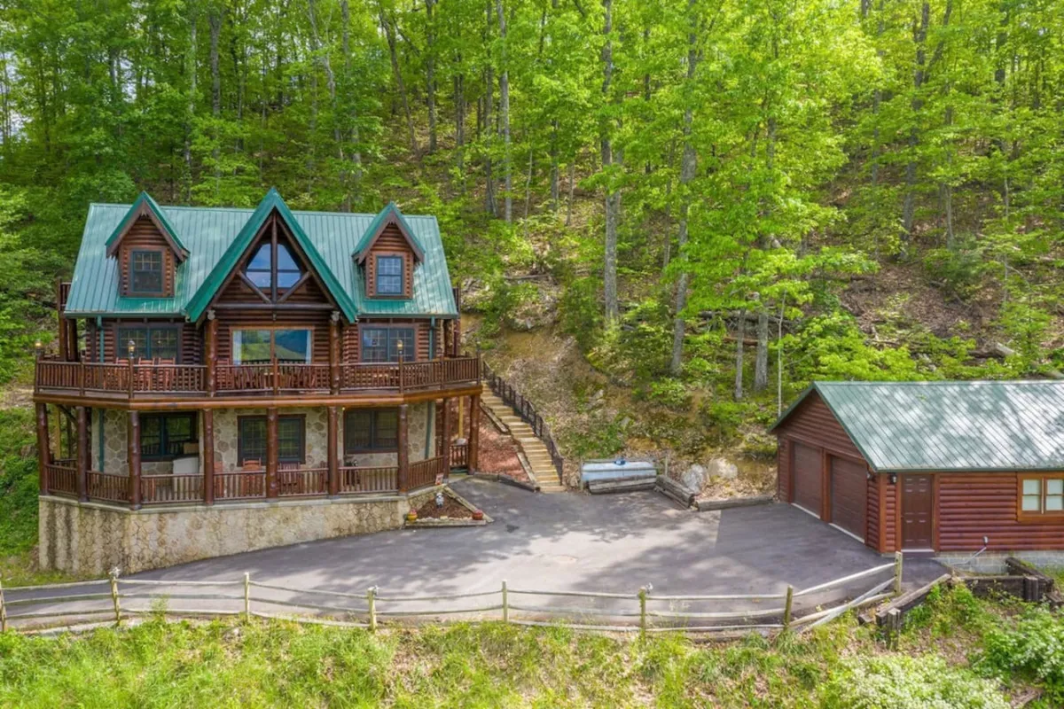 A welcoming view of the cabin's wood exterior, featuring a green roof, stone foundation, and surrounded by lush trees. The spacious driveway and inviting porch set the scene for group arrivals and outdoor gatherings.