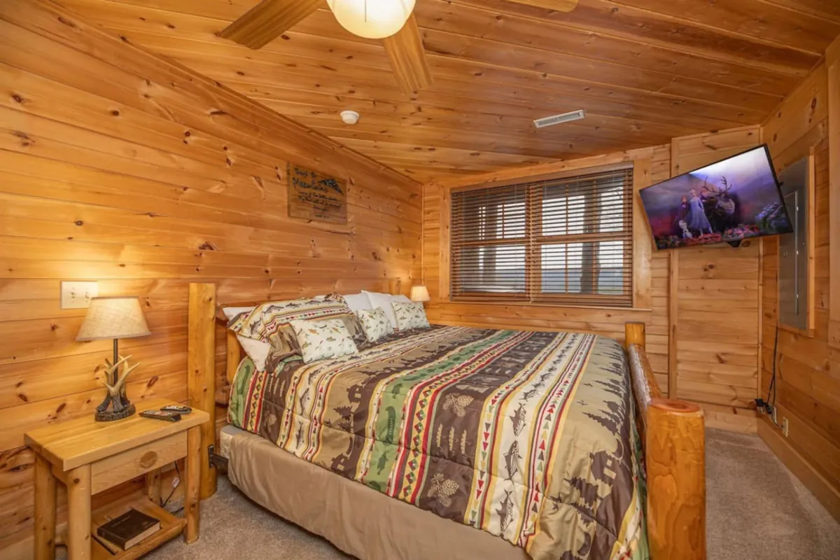 A bedroom featuring wooden walls, a bed with patterned bedding, a nightstand with a lamp, and a mounted television. The room feels both rustic and comfortable, with cozy lighting ideal for unwinding.