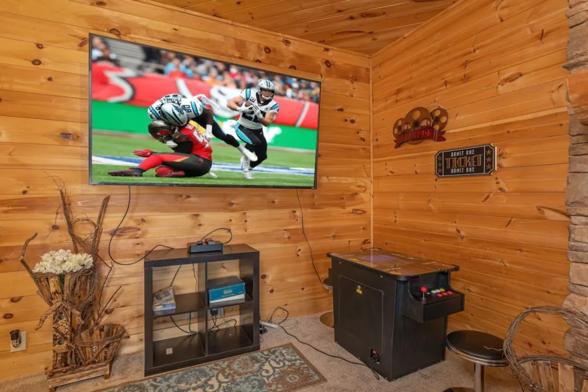 A playful game room corner with a sports game showing on a wall-mounted television and an arcade-style game table below. Wooden walls and tasteful decorative accents add a cozy, cabin-like atmosphere perfect for hours of fun.